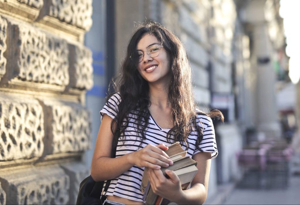 Young woman with a book in hand, smiling as she poses for a photo on campus.