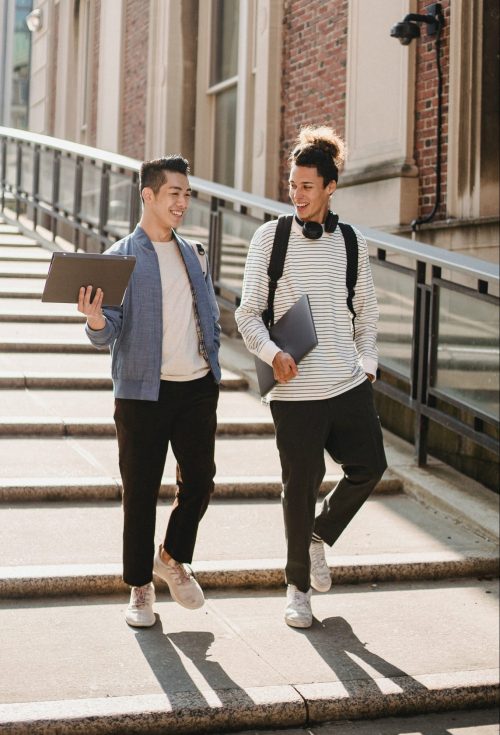 Two young men with books, happily chatting while walking on campus.